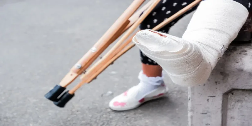 Persona sentada con una pierna enyesada apoyada en el suelo, junto a unas muletas de madera